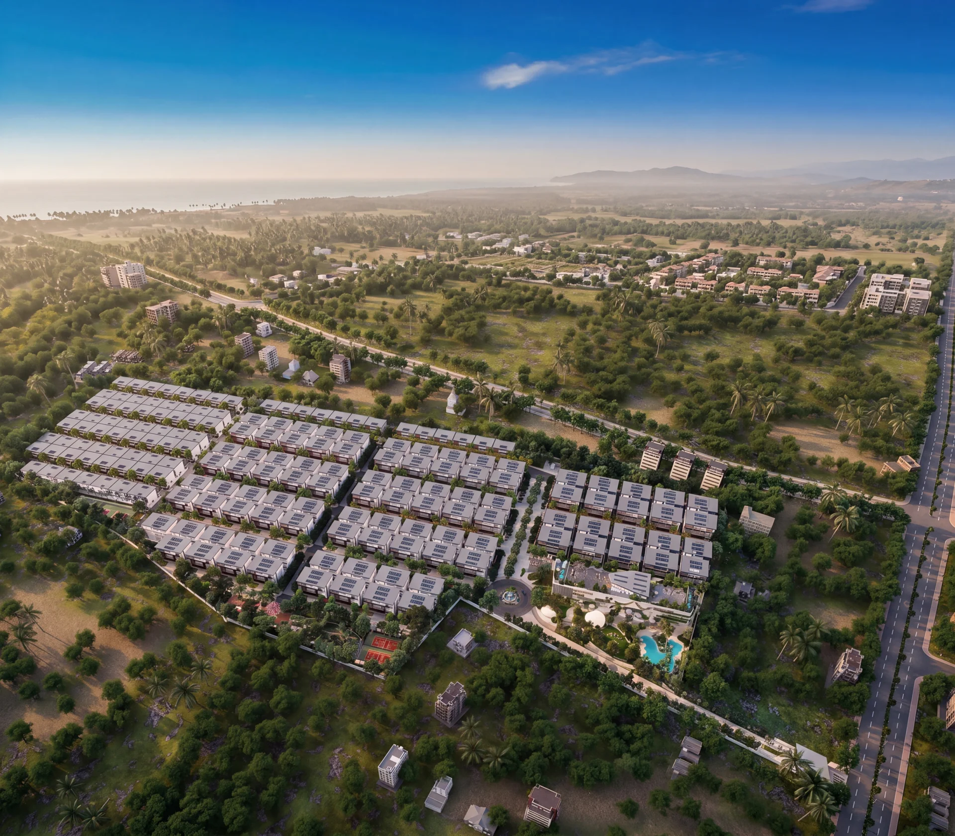 Coastal residential complex with pools, beachfront, roads, and distant mountains under clear blue sky.